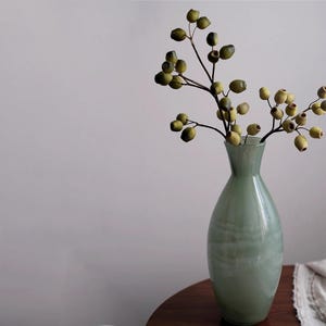 May include: A pale green glass vase holding a branch with olive-green and yellow berries. The vase is on a dark wooden table, with a neutral background. The vase has a rounded shape and a narrow neck, suitable for home decor.
