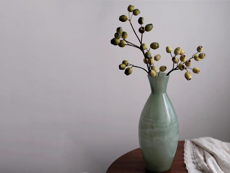 May include: A pale green glass vase holding a branch with olive-green and yellow berries. The vase is on a dark wooden table, with a neutral background. The vase has a rounded shape and a narrow neck, suitable for home decor.