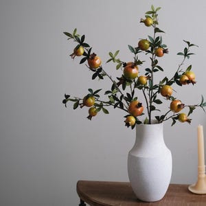May include: A white textured vase holds a decorative arrangement of pomegranate branches. The branches are laden with green leaves and ripe, orange-red pomegranates. A candle and candle holder are on a wooden table.