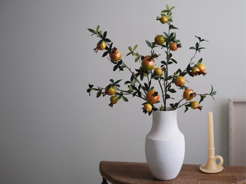 May include: A white textured vase holds a decorative arrangement of pomegranate branches. The branches are laden with green leaves and ripe, orange-red pomegranates. A candle and candle holder are on a wooden table.