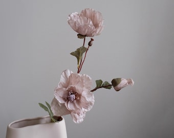May include: A close-up of a floral arrangement featuring light pink poppy flowers in a white vase. The flowers have delicate petals and green leaves. The arrangement is set against a soft gray background.