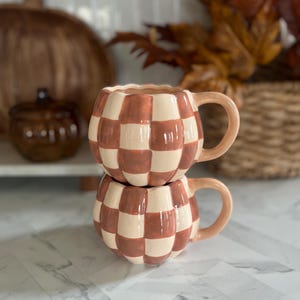 May include: Two stacked ceramic mugs featuring a rounded, pumpkin-like shape with a glossy finish. The mugs are decorated with a repeating checkerboard pattern in alternating shades of cream and terracotta orange, each with a matching curved handle.