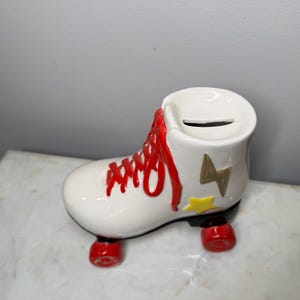 May include: White ceramic roller skate piggy bank with red laces and yellow and gold lightning bolt designs. The skate has red wheels.