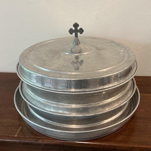 May include: A stack of three silver-colored metal trays, one with a lid, topped with a small cross. The trays are round with a slightly raised edge. The lid has a small cross-shaped handle.