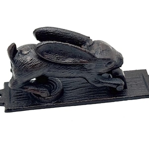 May include: A dark brown, cast iron door knocker shaped like a rabbit in a leaping position. The rabbit is attached to a rectangular mounting plate with screw holes on each end. The metal surface has a textured, rustic finish throughout.