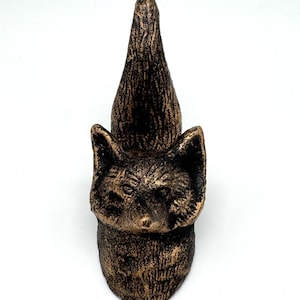 May include: A bronze-colored fox figurine with a textured surface. The fox has pointed ears and a long, pointed head, resembling a gnome. The figurine is set against a white background.