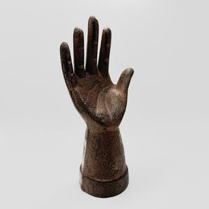 May include: A cast iron hand sculpture with a textured finish. The hand is raised with fingers extended and the palm facing forward. The sculpture is a dark brown color.