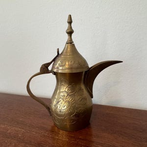 May include: A gold-colored brass teapot with an ornate floral design and a hinged lid. The teapot has a spout and a handle.