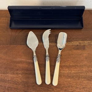 May include: A set of three serving utensils with silver blades and cream-colored handles. The set includes a pie server, a cheese knife, and a spoon. The handles have decorative silver accents. A dark blue box is in the background.