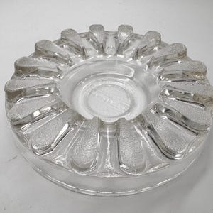 May include: Clear glass ashtray with a circular design. The ashtray features a textured, frosted pattern around the rim and a smooth, circular center. The design includes petal-like shapes around the edge. A vintage item.