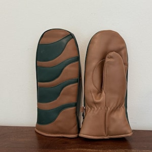 May include: A pair of leather mittens. One mitten features a brown and dark green wavy design, while the other is a solid tan color. The mittens are made of leather and appear to be well-stitched.