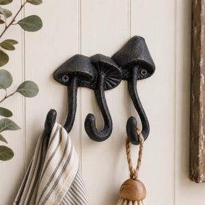 May include: A black cast iron wall hook featuring a trio of mushroom shapes. The mushroom caps serve as the mounting points with visible screws, while the stems curve downward to form three individual hooks for hanging items like towels or brushes.