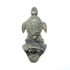May include: A wall-mounted bottle opener shaped like a sea turtle with a distressed, dark bronze and gold-toned finish. The turtle is positioned above the opener mechanism, which features two circular screw holes for mounting to a flat surface.