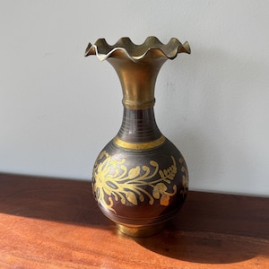 May include: A decorative brass vase with a ruffled rim. The vase has a dark brown body with gold floral designs and a gold neck. The base is also gold. The vase is a decorative item.