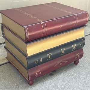 May include: A unique side table designed to resemble a stack of books. The table features a burgundy top, gold-colored pages, and a dark green book in the middle. The table rests on small, rounded feet.