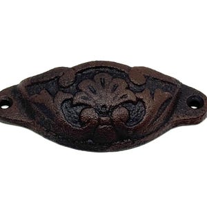 May include: A dark brown, cast iron drawer pull with an ornate floral design. The pull has a curved shape with two holes for mounting. The surface has a textured, aged appearance, suggesting a vintage or antique style.