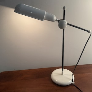 May include: A white and black desk lamp with a long, adjustable arm and a cylindrical shade. The lamp is on a wooden table.