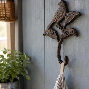 May include: A dark brown, cast iron wall hook featuring a bird perched on a branch with textured leaves. The hook is mounted with two visible screws and holds a white and black striped fabric towel by a small loop at the bottom of the curved hook.