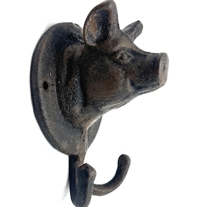 May include: A rustic, cast iron pig head wall hook. The hook is dark brown with a textured finish, featuring a pig's snout, ears, and a curved hook below. The hook is mounted on a circular base, perfect for a farmhouse or country-themed decor.