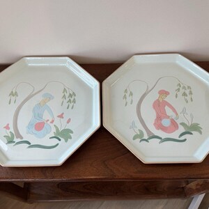 May include: Two octagonal ceramic serving trays with a white background and a painted design of a person in a blue dress and a person in a pink dress standing under a tree with green leaves and pink flowers.