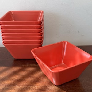 May include: A stack of eight square, coral-colored bowls. One bowl is separate, in front of the stack. The bowls are made of a solid material and have a glossy finish. The bowls are on a dark wooden surface.
