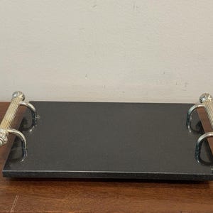 May include: A rectangular black marble serving tray with silver-colored handles. The handles have a decorative, column-like design with ornate detailing on the ends. The tray is displayed on a wooden surface.