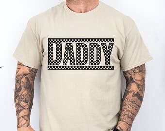 May include: A beige t-shirt with the word "DADDY" in a black checkered design. The text is centered on the front of the shirt. The shirt has short sleeves and a crew neck.