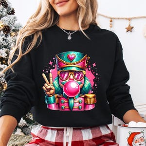 May include: Black sweatshirt featuring a colorful graphic of a nutcracker wearing sunglasses and blowing a bubblegum bubble. The nutcracker is in a military-style uniform with a peace sign. The design includes pink, teal, and gold accents.