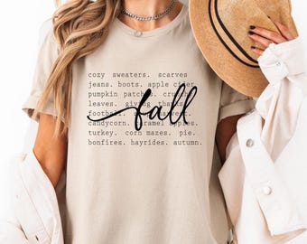 May include: A beige t-shirt with the word "fall" in a cursive font. The shirt features a list of fall-themed words such as "cozy sweaters," "pumpkin patch," and "turkey." The model is wearing a hat and a white jacket.