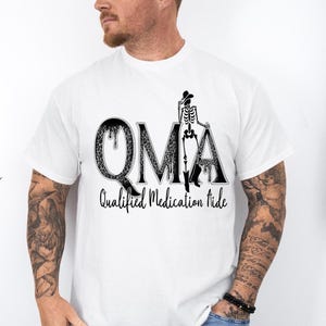 May include: White t-shirt featuring the word "OMA" in a black, patterned font, with a skeleton wearing a cowboy hat and boots. Below the word "OMA" is the text "Qualified Medication Aide" in a black font.