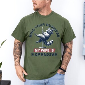 Puede incluir: Camiseta verde oliva con un mapache en una barra con el texto "MIND YOUR BUSINESS" y "MY WIFE IS EXPENSIVE". El mapache es gris y azul. La camiseta es de manga corta.