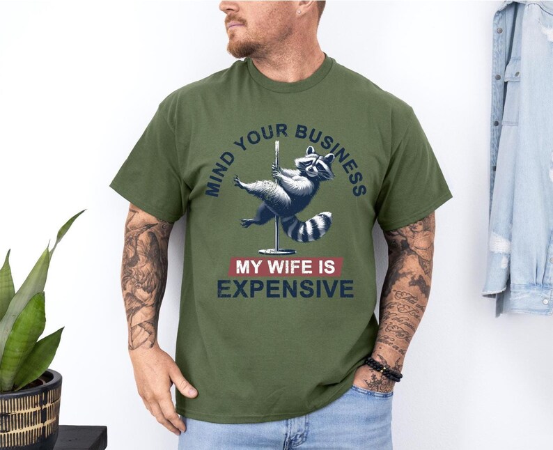 Puede incluir: Camiseta verde oliva con un mapache en una barra con el texto "MIND YOUR BUSINESS" y "MY WIFE IS EXPENSIVE". El mapache es gris y azul. La camiseta es de manga corta.