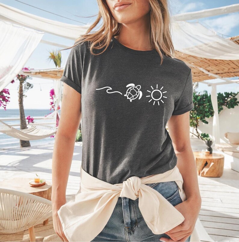May include: Dark gray t-shirt featuring a white graphic of a wave, a turtle, and a sun. The shirt has short sleeves and a relaxed fit. The model is wearing blue jeans and a cream-colored sweater tied around the waist. The background shows a beach scene.