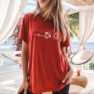 May include: A rust-colored t-shirt featuring a white graphic of a wave, a turtle, and a sun. The short-sleeved shirt is made of a soft material. The design is centered on the front of the shirt.
