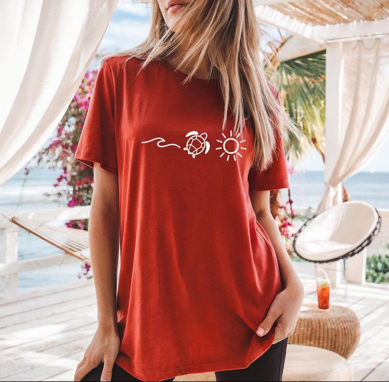 May include: A rust-colored t-shirt featuring a white graphic of a wave, a turtle, and a sun. The short-sleeved shirt is made of a soft material. The design is centered on the front of the shirt.