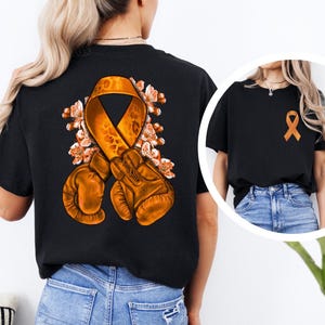 May include: Black t-shirt featuring an orange awareness ribbon, boxing gloves, and floral accents. The design is on the back of the shirt, with a smaller ribbon on the front. The shirt is paired with blue jeans.