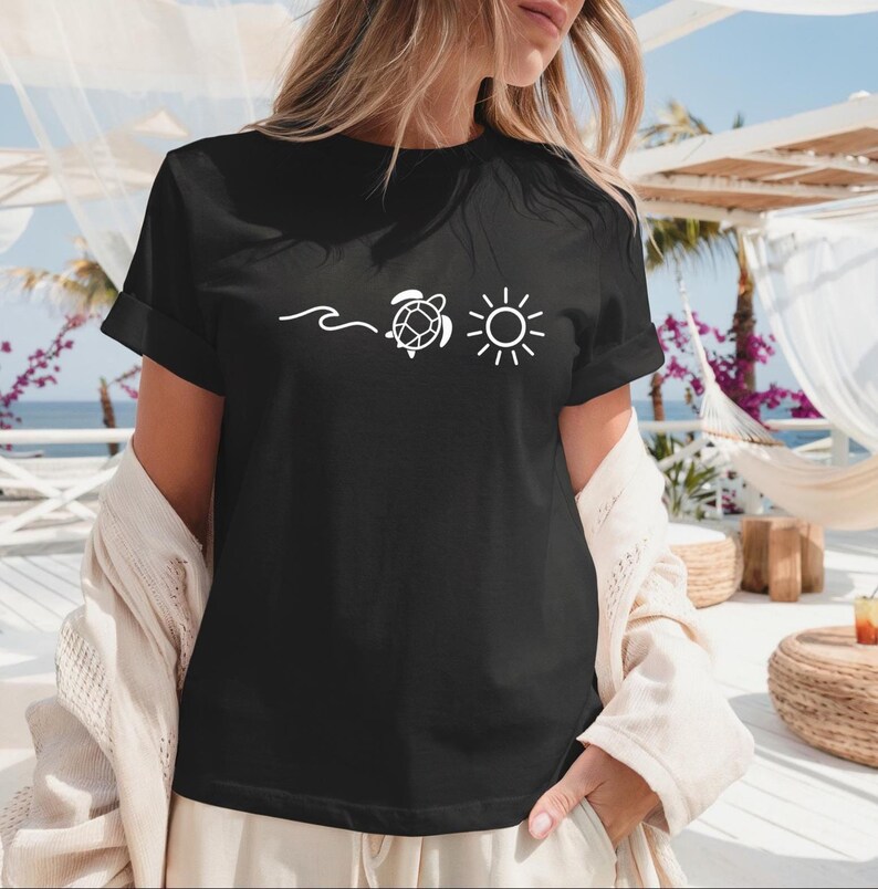 May include: Black t-shirt with a white graphic of a wave, a turtle, and a sun. The shirt has short sleeves and a relaxed fit. The image is taken outdoors, with a beach scene in the background.