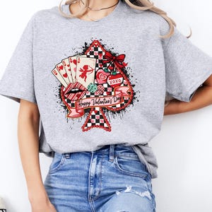 May include: A gray t-shirt featuring a Valentine's Day design. The design includes a spade shape with a red and black checkered pattern, playing cards, a cupid, hearts, and the text "Happy Valentine's Day".