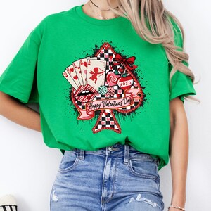 May include: A green t-shirt featuring a Valentine's Day design. The design includes a spade shape with playing cards, hearts, a cupid, and the text "Happy Valentine's Day". The design has red, black, and white accents.