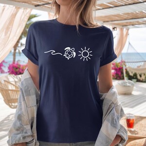 May include: Navy blue t-shirt with a white graphic of a wave, a turtle, and a sun. The shirt has rolled-up sleeves and is made of a soft material. The shirt is part of a beach-themed collection.