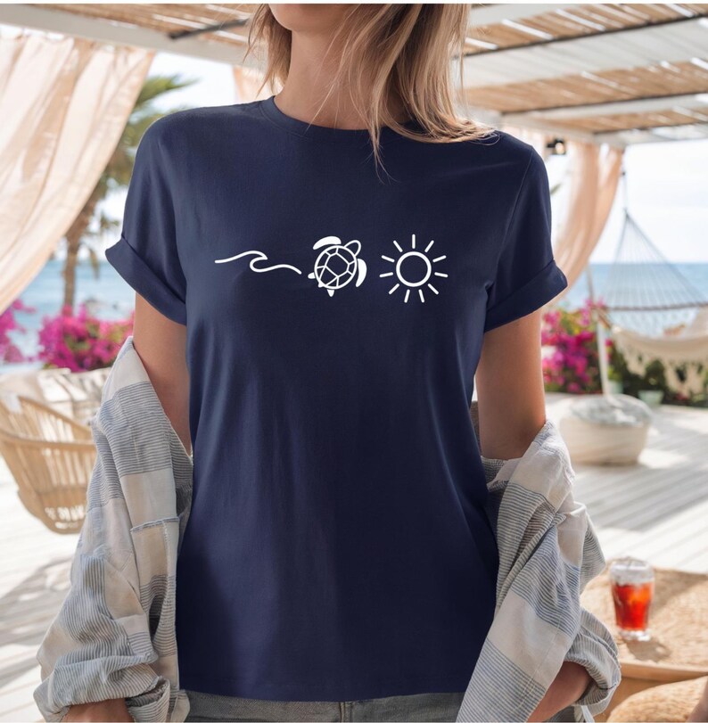 May include: Navy blue t-shirt with a white graphic of a wave, a turtle, and a sun. The shirt has rolled-up sleeves and is made of a soft material. The shirt is part of a beach-themed collection.