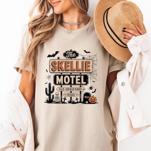 May include: Beige t-shirt featuring a Halloween-themed graphic. The design includes a sign that reads "The Skellie Motel" with a spooky road sign, bats, a spiderweb, a cactus, a skeleton, and a jack-o'-lantern. The shirt has short sleeves.