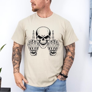 May include: Beige t-shirt featuring a black and white skull graphic. The skull is depicted with bony hands extending from each side, both displaying the middle finger. The design is centered on the shirt.
