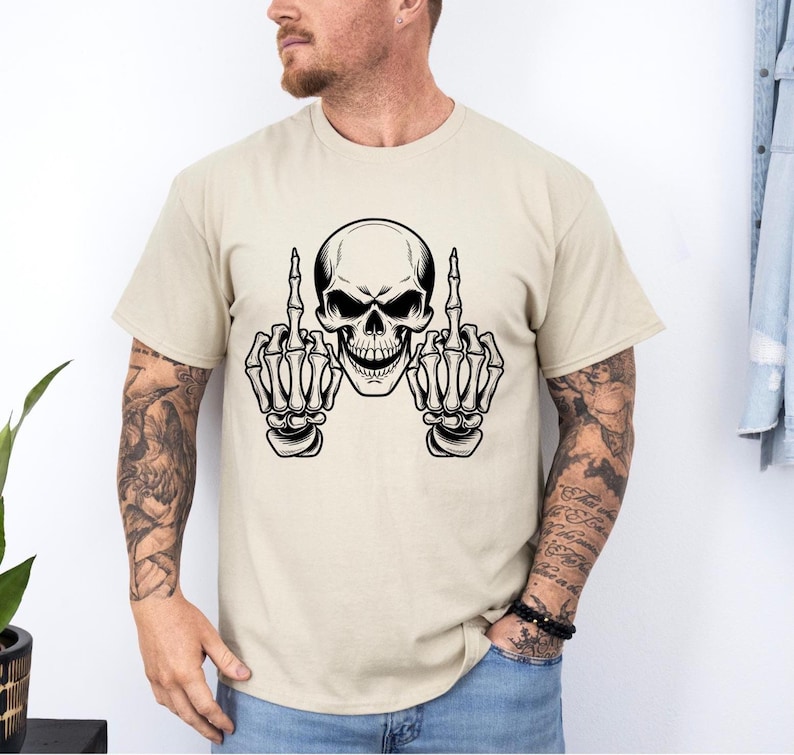 May include: Beige t-shirt featuring a black and white skull graphic. The skull is depicted with bony hands extending from each side, both displaying the middle finger. The design is centered on the shirt.