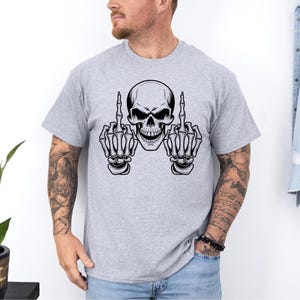 May include: A light gray t-shirt featuring a black and white skull graphic. The skull is depicted with its hands up, displaying the middle finger on each hand. The design is centered on the shirt.
