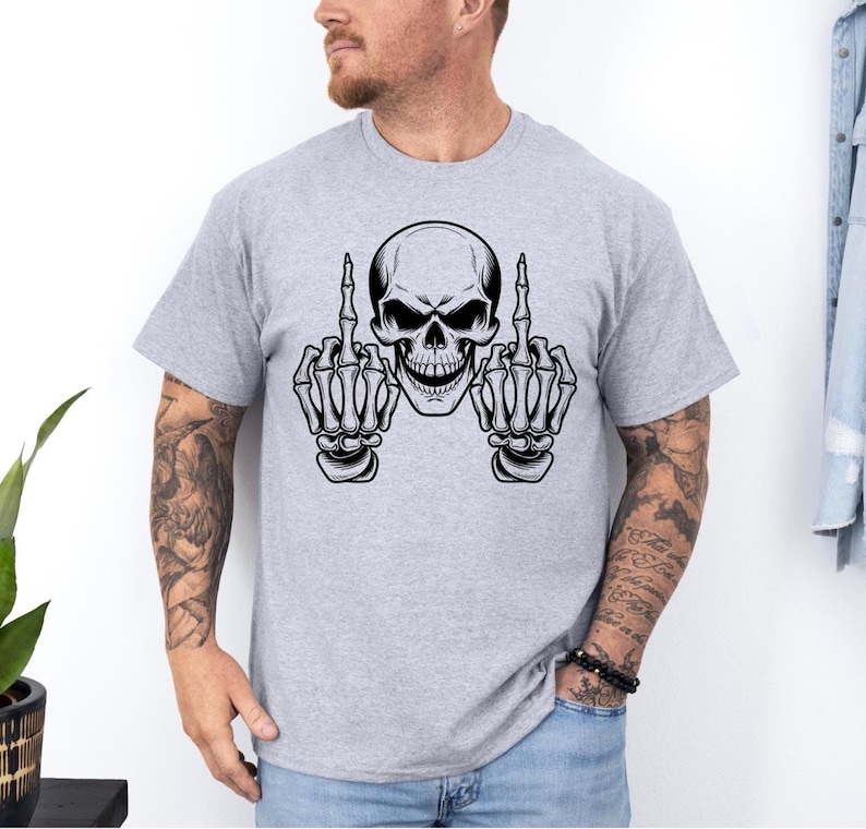 May include: A light gray t-shirt featuring a black and white skull graphic. The skull is depicted with its hands up, displaying the middle finger on each hand. The design is centered on the shirt.