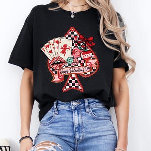May include: Black t-shirt featuring a Valentine's Day design. The design includes a spade shape with playing cards, lips, hearts, and a bow. The words "Happy Valentine's Day" are written on a banner.