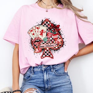 May include: A light pink t-shirt featuring a Valentine's Day graphic. The graphic includes playing cards, hearts, lips, and the text "Happy Valentine's Day" on a spade shape. The design incorporates red, black, and white colors.