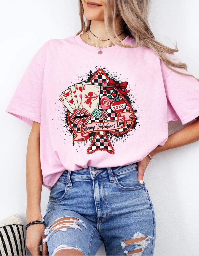 May include: A light pink t-shirt featuring a Valentine's Day graphic. The graphic includes playing cards, hearts, lips, and the text "Happy Valentine's Day" on a spade shape. The design incorporates red, black, and white colors.
