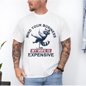 Puede incluir: Camiseta blanca con un mapache en una barra con el texto "MIND YOUR BUSINESS" arriba. Debajo, el texto "MY WIFE IS EXPENSIVE". Es una prenda casual, adecuada para el uso diario.